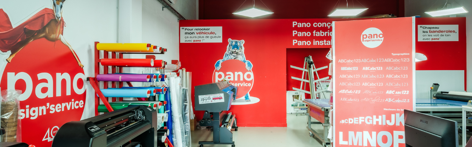Intérieur agence PANO avec racks d'adhésifs, plotter de coupe, traceur numérique et mur orné des logos PANO